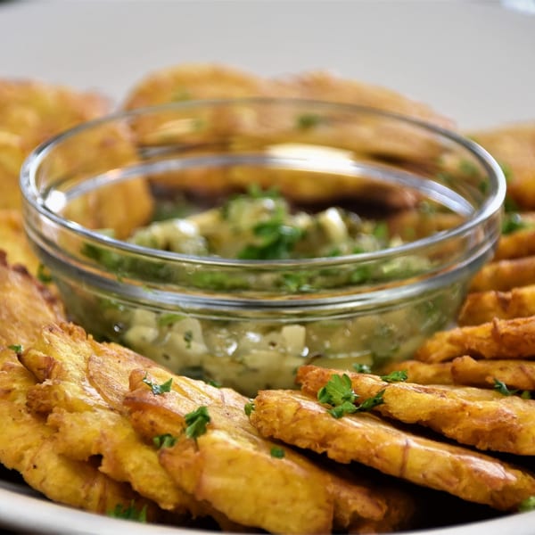 Golden-brown fried plantains, or tostones, arranged around a glass bowl of green dipping sauce.