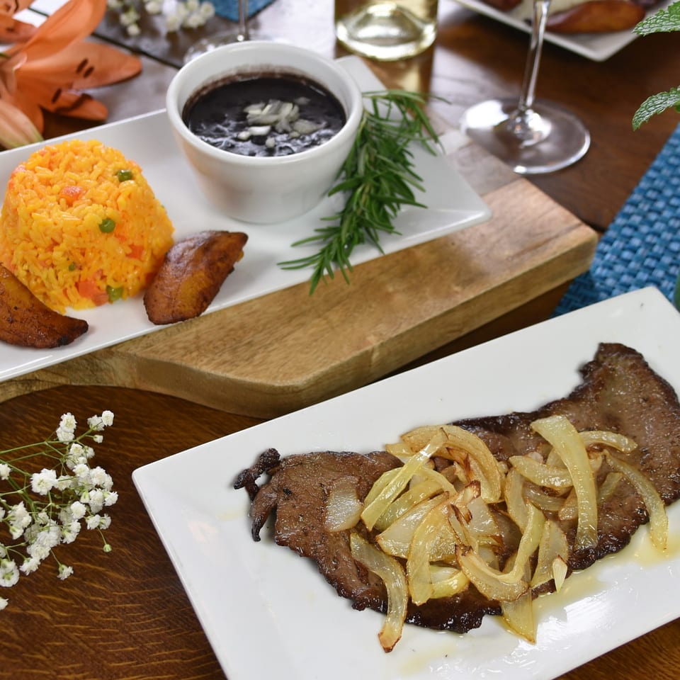 Plates of grilled steak with sautéed onions, orange rice, fried plantains, and black beans on a wooden dining table.