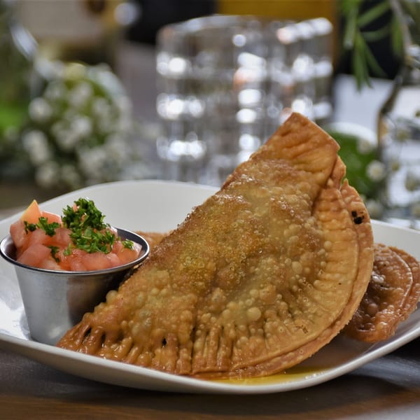 Two golden-brown Cuban empanadas with a side of diced tomatoes and parsley on a white plate.