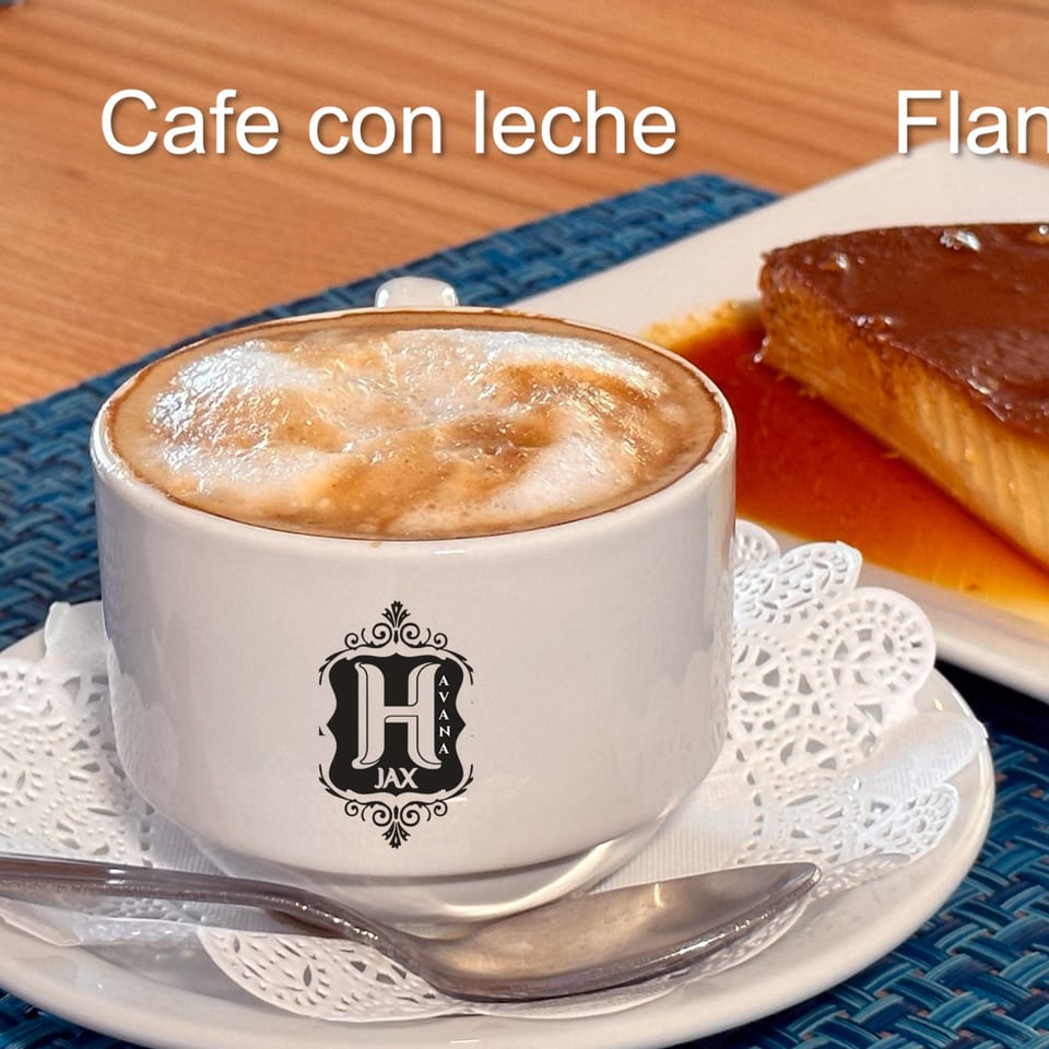 Café con leche in a branded white cup served with flan on a plate, spoon, and decorative doily.