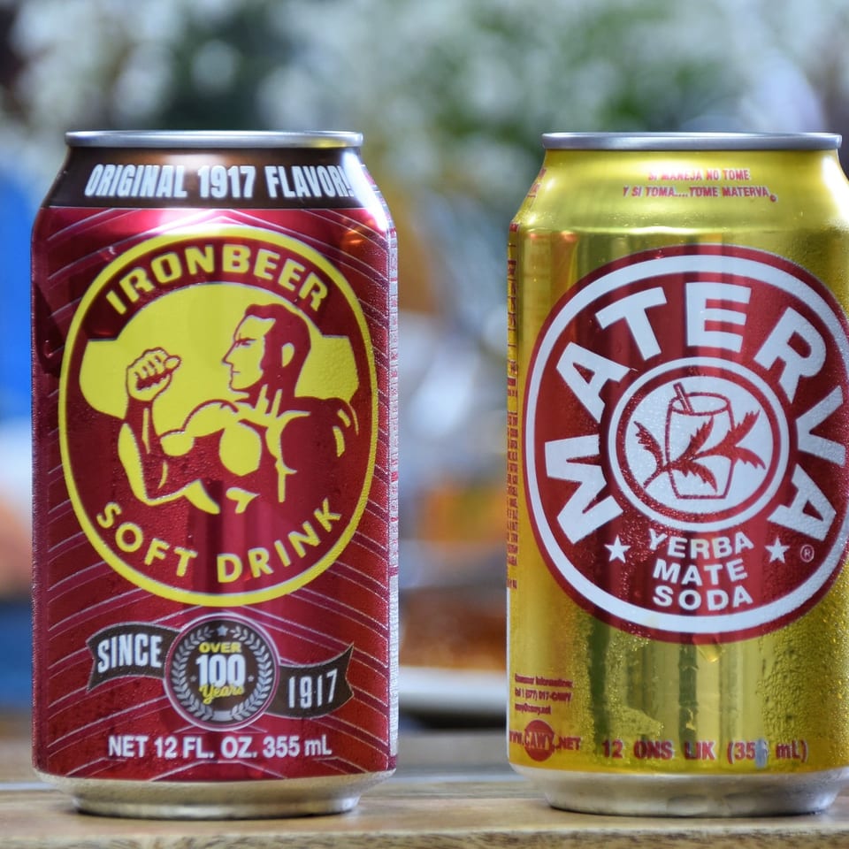 Two cans of Ironbeer soft drinks sit on a wooden surface, showing red original flavor and yellow yerba mate soda branding.