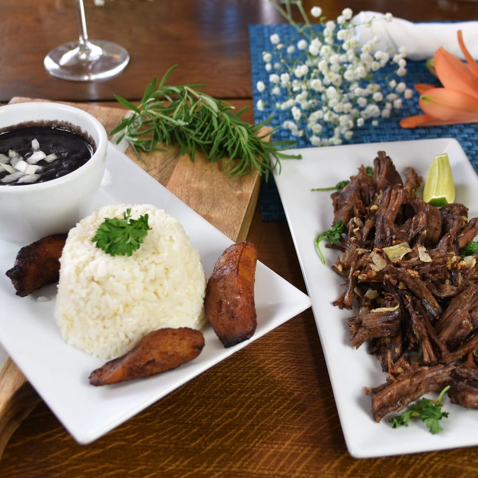 Plates of white rice, fried plantains, black beans, and crispy fried meat garnished with herbs and lime.