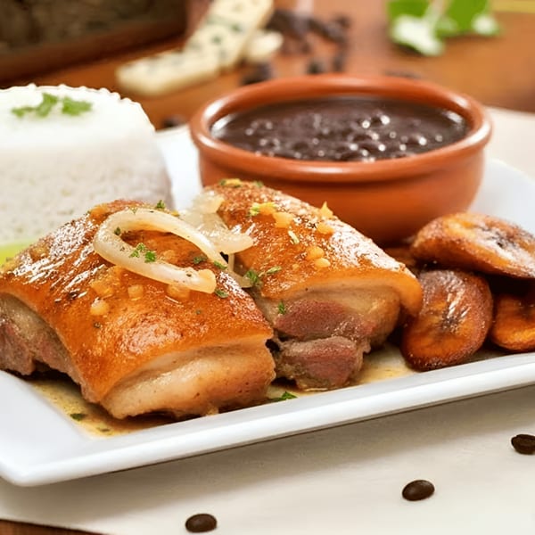 Cuban dish with crispy pork belly, white rice, black beans, and fried plantains on a white plate.