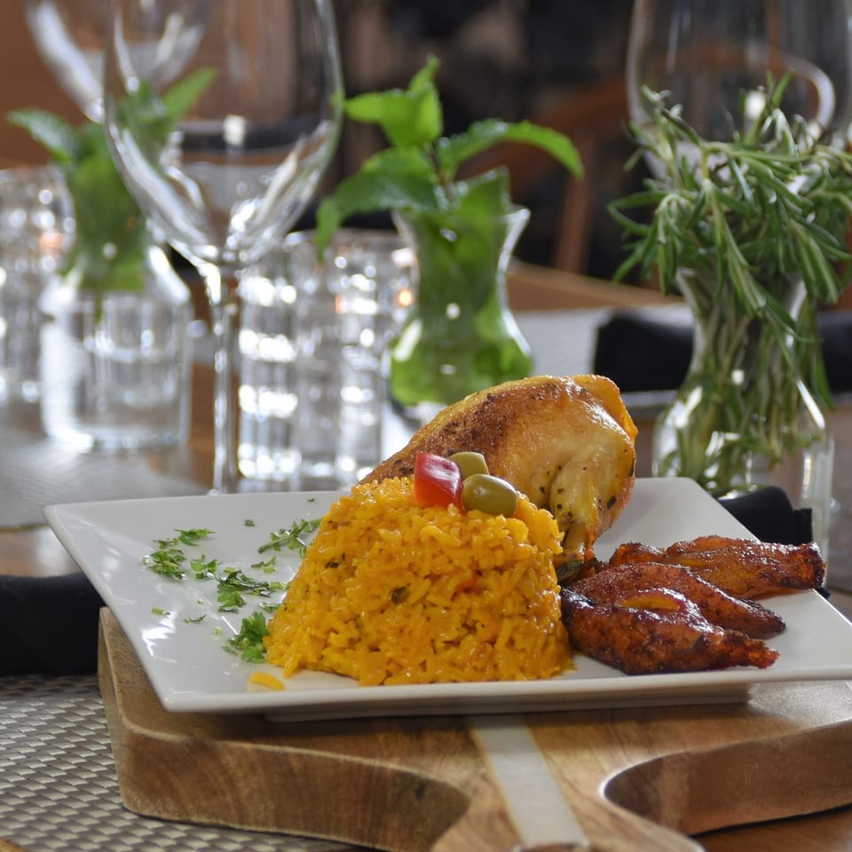 Plated grilled chicken with golden rice, roasted vegetables, herbs, and olives on a white square dish at a restaurant table.