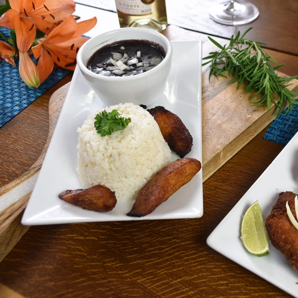 White rice with grilled sausages and dark bean stew garnished with onion, served on a white plate beside rosemary and orange lilies.